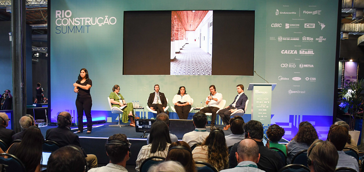 Auditório lotado em primeiro plano com cinco debatedores no palco e um telão ao fundo mostrando o patrimônio histórico de Petrópolis
